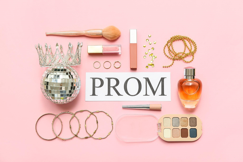 A paper with the word prom written in capital letters is placed on a pink background with a disco ball and prom accessories surrounding it